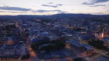 Şehir merkezindeki binaların üzerinden geriye doğru uçuyor. Gökyüzünün alacakaranlık rengine karşı şehir manzarası. Şehir körfezinde su yüzeyi açığa çıkıyor. Oslo, Norveç.
