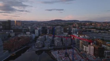 Şehir merkezindeki modern çok katlı binaların üzerinde renkli alacakaranlık gökyüzüne karşı ilerliyorlar. İnşaat alanında kule vinçleri. Oslo, Norveç.