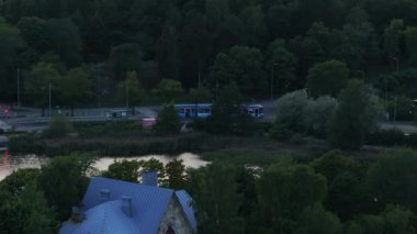 Şehrin dışındaki caddelerde arabaların yanından geçen tramvayın hava görüntüsü. Gün batımından sonra parklarda ağaçlar. Toplu taşıma. Helsinki, Finlandiya.