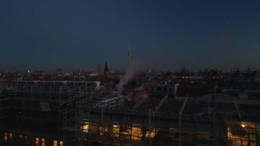 Hava kararınca şehir merkezindeki binaların üzerinden ilerliyorlar. Çok katlı evin etrafında iskeleler var. Duman ya da duman çatıdan yükseliyor. Uzun Fernsehturm 'la akşam manzarası. Berlin, Almanya.