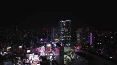 Geceleri şehrin üzerinden geriye doğru uçuyor. Şehir merkezindeki binalar ve sokaklar. Aydınlatılmış ticari merkez ve büyük video ekranları. Tokyo, Japonya.