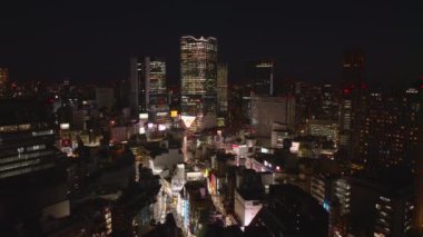 Gece şehrinin üzerinde geriye doğru uç. Shibuya bölgesinde sokaklar ve binalar aydınlatıldı. Modern yüksek binalar. Tokyo, Japonya.