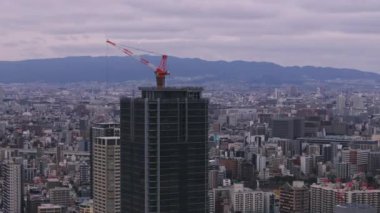 Kırmızı ve beyaz çizgili vinçli inşaat alanının havadan kayma ve pan görüntüleri. Şehirde konut ya da ofis kulesi var. Osaka, Japonya.