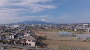 İleriye doğru tren hattının ve kasabadaki binaların üzerinden uçar. Nehir kıyısındaki büyük seralar. Uzakta Fuji Dağı tepesi olan ikonik dağlar. Japonya.