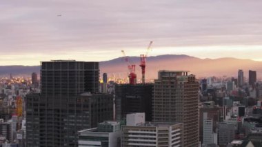Metropolis 'teki binaların havadan görüntüsü. Yüksek binaların tepesinde bir çift kırmızı ve beyaz çizgili vinç. Yükselen güneş bulutların arasında parlıyor. Osaka, Japonya.