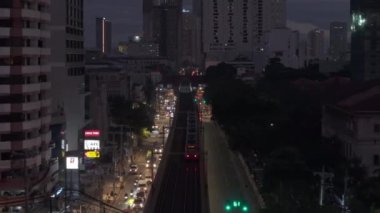 Geniş caddenin üzerinde, ortada demiryolu ile ilerliyorlar. Yolcu treni akşam kentinde yüksek bir hat üzerinde gidiyor. Manila, Filipinler.