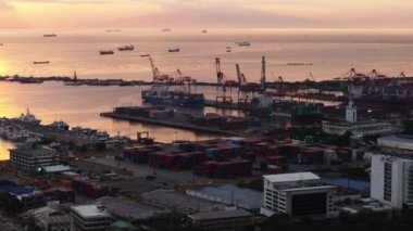 Büyük vinçlerle ve deniz aşırı konteynırlarla dolu kargo deniz limanının havadan görüntüsü. Kıyıda bekleyen gemiler, renkli günbatımına karşı manzara. Manila, Filipinler.