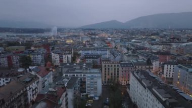 Akşam karanlığında şehrin havadan panoramik manzarası. Şehir merkezindeki çok katlı binaların üzerinden uçuyor. Dağlar ve arka planda ünlü Jet d Eau çeşmesi. Cenevre, İsviçre.