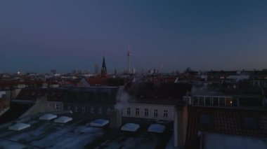 Alacakaranlıkta şehir merkezindeki çatıların üzerinde alçak uçuş. Kilise kulesi ve Berliner Fernsehturm gökyüzüne karşı. Berlin, Almanya.