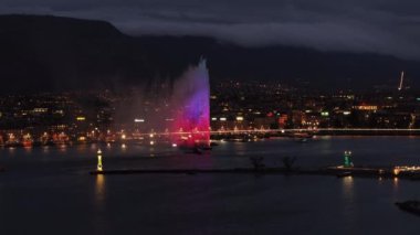 Genevas jet deau fıskiyesi gece aydınlandı, şehir manzarasına ve Lac Leman suyuna canlı renkler dökülüyor.