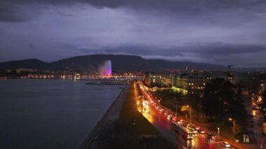 Genevas rıhtımının ve jet deau 'nun karanlık çöktüğünde çekilmiş insansız hava aracı görüntüleri, manzaralı bir arka planda yoğun trafiği gösteriyor..