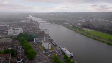 Meuse nehri ile Maastricht şehir manzarası, bulutlu gökyüzü altında binalar, altyapı ve yeşil alanlar sergileniyor..