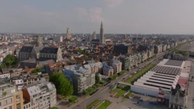 Antwerps canlı şehir merkezinde kalabalık caddelerde hareket eden arabalar ve tramvaylar, ikonik mimari ve arka planda çarpıcı bir katedral kulesi yer alıyor..