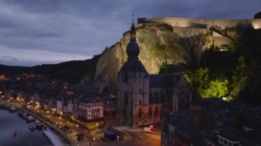 Havadan bakıldığında, aydınlanmış notre de Dinant kolej kilisesi ve alacakaranlıktaki Meuse nehri boyunca canlı şehir merkezi görülüyor..