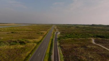 Yeşil bitki örtüsüyle çevrili düz bir yol üzerinde ilerleyen hava manzarası ve açık bulutlu mavi gökyüzünün altında halkalı fiyort..