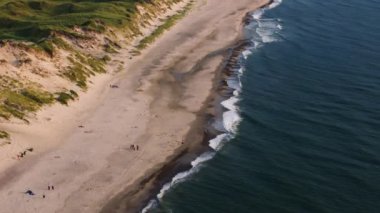 Turkuaz dalgalar, Ringkobing Fjord 'daki çimenli kum tepelerinin altında kumlu bir sahilin kıyısına doğru akıyor. Turistler sahil boyunca yürür..