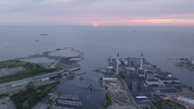 Hava görüntülerinde Kopenhag 'ın endüstriyel limanı gün batımının sıcak renkleriyle aydınlatılıyor. Uzak bir rüzgar çiftliği, yenilenebilir enerji kaynaklarına ithaf edilen şehirleri vurguluyor.