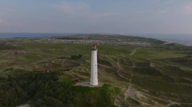 White lighthouse is standing on a sandy coastline surrounded by green vegetation. The North Sea is visible in the background.