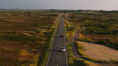 Düz, çimenli, çalılık bir arazide, yanında bisiklet yolu olan kaldırımlı bir yol boyunca giden arabaların hava manzarası..