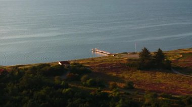 Danimarka 'daki Kuzey Denizi kıyı şeridini gösteren hava görüntülerinde, parlak güneş ışığı altında yemyeşil bitki örtüsü ve sakin sularla çevrili bir rıhtım ve küçük bir bina yer alıyor..