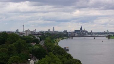 Panoramik manzara, bulutlu bir gökyüzü altında yemyeşil ağaçlar tarafından çerçevelenmiş Ren Nehri ve Hohenzollern Köprüsü 'nün yer aldığı Colognes siluetini yakalar..