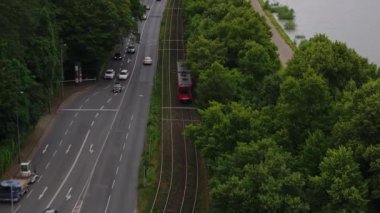 Almanya 'nın Köln banliyölerinden geçen kırmızı bir tramvayın hava görüntüsü. Yol boyunca yemyeşil ağaçlar var ve Ren Nehri sokağın kenarında akıyor..
