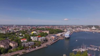 Finlandiya, Helsinki 'nin hava görüntülerinde güneşli bir yaz gününde limana yanaşan büyük bir yolcu gemisi görülüyor ve canlı şehir manzarası ve hareketli liman manzarası gösteriliyor..