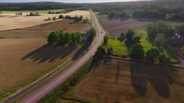 Havadan bakıldığında, Finlandiya 'da yemyeşil tarlalar ve sık ormanlarla çevrili, kırsal bir yol boyunca giden siyah bir araba görülüyor. Sahne huzurlu bir yaz manzarası yakalar..