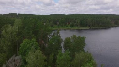 Havadan bakıldığında, yemyeşil ormanlarla çevrili sakin bir göl bulunan çarpıcı bir İsveç manzarası görülüyor. Kamera zarifçe sağa hareket ediyor, bölgenin doğal güzelliğini vurguluyor..