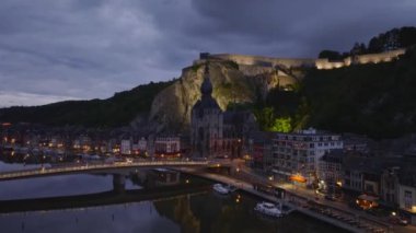 Arabalar Dinant, Belçika 'da bir köprüden geçiyor. Kale, alacakaranlıkta Meuse nehrinin üzerindeki uçurumda aydınlanır..