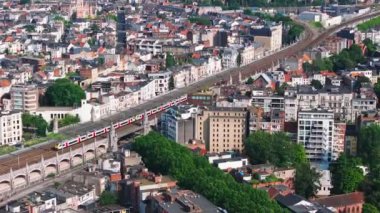 Kırmızı ve beyaz tren, Belçika 'nın Antwerp şehrinde demiryolu boyunca yerleşim binaları ve yemyeşil ağaçlarla çevrili bir köprüden geçer..