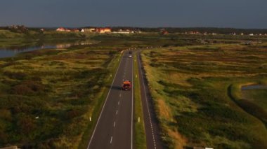 Danimarka 'da Ringkobing Fjord yakınlarındaki bir kır yolunda ilerleyen turuncu kamyon, uzaktan görülebilen Kuzey Denizi, günbatımında manzaralı bir yaz manzarası yaratıyor..