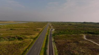 Yeşil bitki örtüsüyle çevrili düz bir yol üzerinde ilerleyen hava manzarası ve açık bulutlu mavi gökyüzünün altında halkalı fiyort..