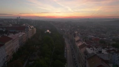 Güneş doğarken Prag şehrinin havadan görünüşü aşağıdaki binaların ve sokakların üzerine sıcak bir ışık saçıyor. Arabalar yemyeşil bir parkın sınırında yol boyunca ilerliyor..