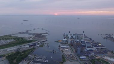 Hava görüntülerinde Kopenhag 'ın endüstriyel limanı gün batımının sıcak renkleriyle aydınlatılıyor. Uzak bir rüzgar çiftliği, yenilenebilir enerji kaynaklarına ithaf edilen şehirleri vurguluyor.