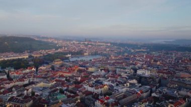 Güneş ışığı Prag şehir merkezindeki binaları aydınlatır, ortasından akan bir nehir ve uzaktaki tepeler.