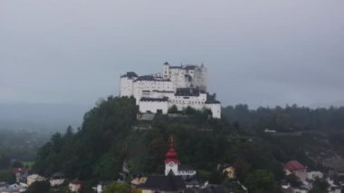 Avusturya, Salzburg 'daki Hohensalzburg Kalesi, yağmurlu bir günde dramatik bulutların olduğu bir tepenin üzerinden görkemli bir şekilde yükseliyor..