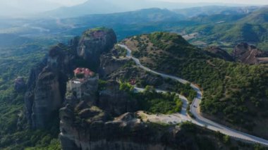 Hava perspektifleri, bir Meteora kaya oluşumunun tepesindeki bir manastırın yanındaki yolda dönen arabaların, UNESCO Dünya Mirası 'nın nefes kesen manzarasını gözler önüne serdiğini gösteriyor..