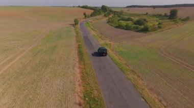 Hava görüntüleri, kırsal yaz yolculuklarının cazibesini gözler önüne seren, canlı tarım alanlarıyla çevrili, dolambaçlı bir kır yolu boyunca seyahat eden siyah bir SUV yakalıyor..