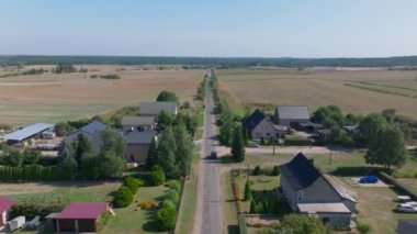 Havadan bakıldığında düz bir yolda seyreden parlak siyah bir araba görülüyor. Canlı tarlalar ve bir köydeki antika evler, çarpıcı bir kırsal kesim manzarası yakalıyor..