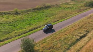 Siyah SUV, yemyeşil çayırlarla çevrili asfalt bir yol boyunca gidiyor. Sahne, kırsal alanların özünü yansıtıyor, doğanın güzelliğini ve sükunetini gösteriyor..