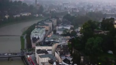 Salzburg 'un eski şehri, içinden Salzach Nehri akan havadan görünüyor. Yayalar nehir kıyısında yürüyorlar ve arabalar caddelerde ilerliyorlar..