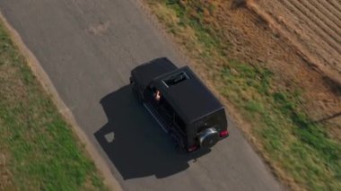 Kırsal alanda asfalt yol boyunca giden siyah bir SUV 'nin hava görüntüsü. Güneşli bir günde yolculuğun tadını çıkaran bir yolcumuz var..