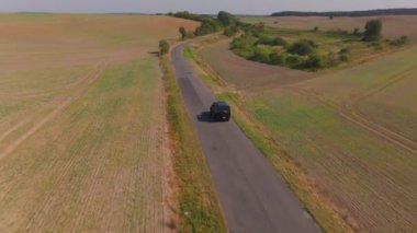 Hava görüntüleri, kırsal yaz yolculuklarının cazibesini gözler önüne seren, canlı tarım alanlarıyla çevrili, dolambaçlı bir kır yolu boyunca seyahat eden siyah bir SUV yakalıyor..