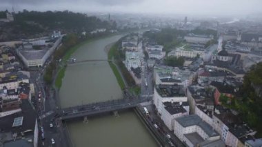 Salzburg şehri, şehir manzarası boyunca akan Salzach Nehri 'ni gözler önüne seriyor. Bulutlu bir gökyüzü şehrin üzerinde kasvetli bir atmosfer yaratıyor..