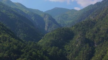Havadan bakıldığında, Yunanistan 'ın Olimpos Dağı' nda yuva yapmış canlı yeşil bir vadi görülüyor. Yoğun ormanlar, açık mavi gökyüzünün altında huzurlu bir atmosferi teşvik ederek araziyi kaplıyor..