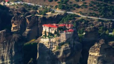 Yunanistan 'ın Meteora kentinde yükselen bir kaya oluşumunun tepesine tünemiş bir manastırın hava görüntüsü, güneş doğada gölgeler oluştururken.