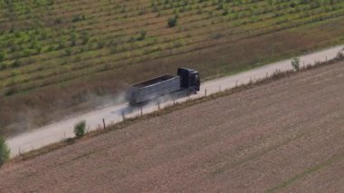 Hava görüntülerine göre tozlu bir kır yolunda yol alan bir kamyon, resimli tarlalardan ve canlı kırsal alanlardan geçerken bir toz bulutunu tekmeliyor..