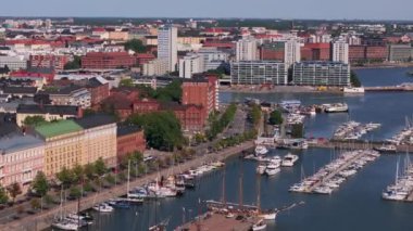 Hava manzarası Helsinki limanını gösteriyor, yat limanına yanaşmış yelkenliler ve turistler canlı bir şehir ortamında, güneşli bir mavi gökyüzünün altında rıhtımda geziniyorlar..