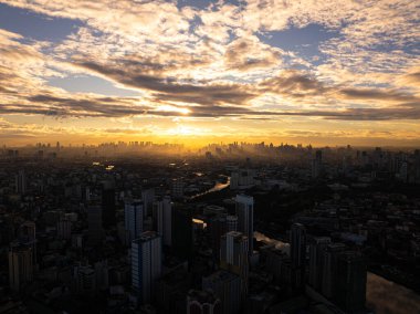 Manila şehrinin gökdelenleri ve binaları bulutlar ve güneş ışınlarıyla altın gün batımında parlıyordu. Bir Asya metropolünün hava görüntüsü..